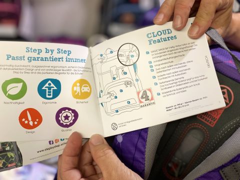 Una persona tiene in mano un opuscolo aperto con simboli colorati per sostenibilità, ergonomia, sicurezza, design e qualità. Sul lato destro sono descritte in dettaglio le "Funzionalità CLOUD" con un diagramma. Sullo sfondo si vede uno zaino.