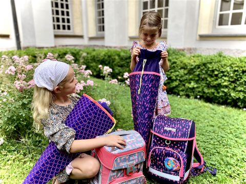Due giovani ragazze sono sedute su un prato in un giardino e ispezionano i loro zaini colorati. Una indossa una fascia per capelli e tiene in mano un materassino isolante viola, l'altra guarda nel suo zaino rosa-viola. Sullo sfondo si vedono cespugli di fiori e finestre.