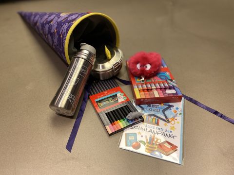 Un grande cono decorato con un motivo viola è pieno di materiale scolastico, tra cui pastelli, una borraccia di metallo, un peluche rosso e biglietti d'auguri. Il cono e gli oggetti sono disposti su una superficie piana.