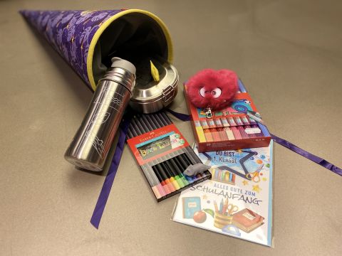Un grande e colorato cono scolastico è aperto e rivela il contenuto, come una borraccia di metallo, pastelli colorati, un peluche rosa e un libro con testo in tedesco. Il cono è decorato con stelle e rivestito con un tessuto viola.