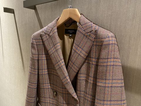 Un blazer a quadri marrone e beige è appeso a una gruccia di legno davanti a una parete beige strutturata. Il blazer presenta un classico motivo a quadri con chiusura a bottoni e un taglio sartoriale.
