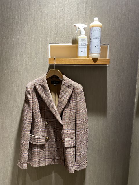 Un blazer a quadri è appeso a una gruccia di legno su una parete strutturata. Sopra c'è una piccola mensola con un flacone spray e una soluzione detergente.