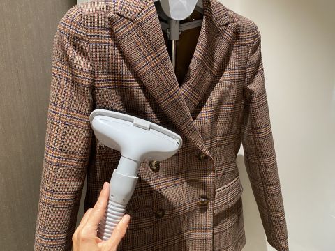 Una mano tiene un ferro da stiro a vapore e lo preme contro un blazer doppiopetto a quadri appeso a un appendiabiti grigio. Il blazer presenta un classico motivo a quadri nei toni del marrone. La scena è ambientata in un interno.