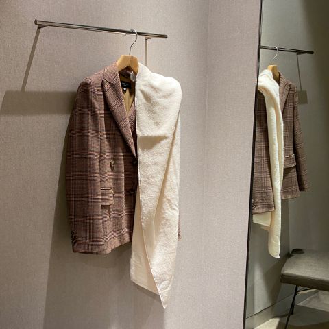 Un blazer a quadri marrone con una sciarpa color crema è appeso a un appendiabiti in un camerino. Il suo riflesso si vede nello specchio e nell'angolo c'è uno sgabello.