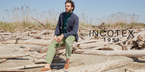 Un uomo è seduto su un legno galleggiante su una spiaggia. Indossa un blazer blu navy, un maglione verde e pantaloni verdi. Abbina scarpe marroni e bianche. Sullo sfondo si vedono erba alta e un cielo azzurro limpido. A destra c'è il testo "INCOTEX 1951".