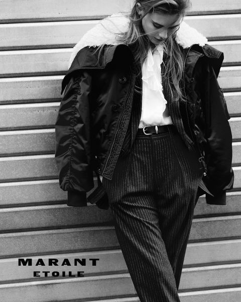 Foto in bianco e nero di una donna che indossa una giacca con colletto in shearling, una blusa con volant e pantaloni gessati, appoggiata a un muro di metallo. Il testo "MARANT ETOILE" è in basso a sinistra.