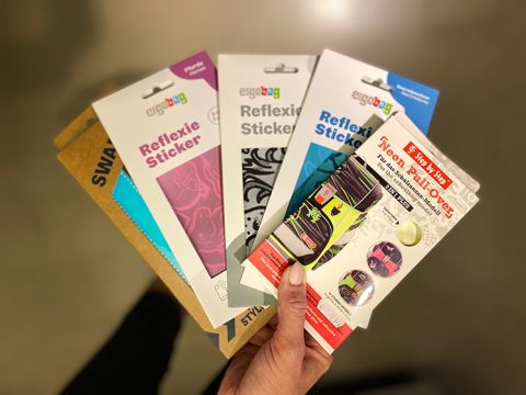 Una mano tiene cinque confezioni di adesivi riflettenti e neon Ergobag in vari colori e motivi, tra cui design rosa, blu e multicolore. Lo sfondo è un interno sfocato.