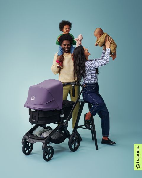 Una gioiosa scena familiare con un uomo che porta un bambino sulle spalle e una donna che solleva un altro bambino. Nelle vicinanze, un passeggino viola si trova di fronte a uno sfondo blu-verde. In basso a destra si vede il logo Bugaboo.