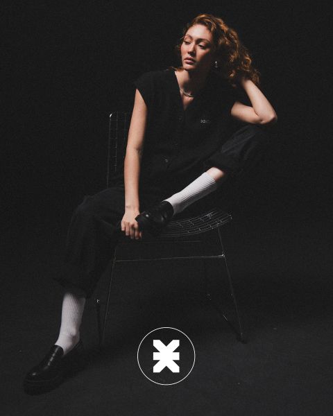 Una persona con i capelli ricci è seduta su una sedia di filo metallico in una stanza buia, indossa abiti neri, calzini bianchi e scarpe basse nere. Una gamba è piegata, un gomito è appoggiato sul ginocchio e sul pavimento c'è un logo circolare con una "X".