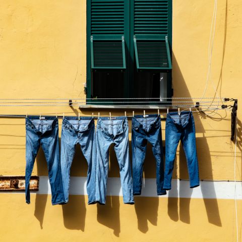 Cinque paia di blue jeans sono appesi a uno stendibiancheria davanti a un muro giallo. Sopra, una finestra con persiane verdi è parzialmente aperta e la luce del sole proietta ombre sul muro.