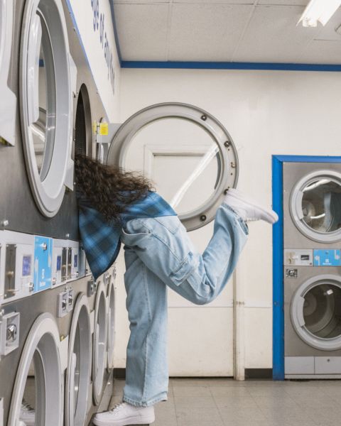 Una persona con una camicia a quadri blu e jeans è appoggiata a una lavatrice in una lavanderia a gettoni. La sua gamba destra è sollevata all'indietro per bilanciarsi. La stanza ha diverse lavatrici e asciugatrici in tonalità blu e bianco.
