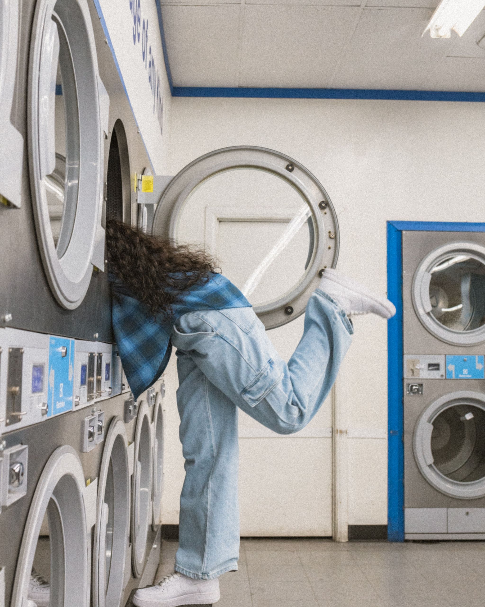 Una persona con una camicia a quadri blu e jeans è appoggiata a una lavatrice in una lavanderia a gettoni. La sua gamba destra è sollevata all'indietro per bilanciarsi. La stanza ha diverse lavatrici e asciugatrici in tonalità blu e bianco.