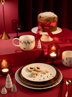 Una tavola imbandita a festa con una grande torta, avvolta in un nastro rosso e cosparsa di zucchero a velo, su un'alzata. Ci sono biscotti sui piatti, tazze a tema, candele, decorazioni natalizie rosse e bianche e uno sfondo rosso intenso.