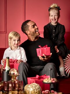 Un uomo siede con una scatola regalo rossa su un divano rosso e sorride a due bambini piccoli. Un bambino, vestito di bianco, tiene in mano un regalo rosso simile, mentre l'altro, vestito di nero, è seduto sullo schienale del divano. In primo piano c'è un tavolo con dolcetti e una bottiglia.