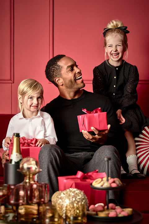 Un uomo siede con una scatola regalo rossa su un divano rosso e sorride a due bambini piccoli. Un bambino, vestito di bianco, tiene in mano un regalo rosso simile, mentre l'altro, vestito di nero, è seduto sullo schienale del divano. In primo piano c'è un tavolo con dolcetti e una bottiglia.