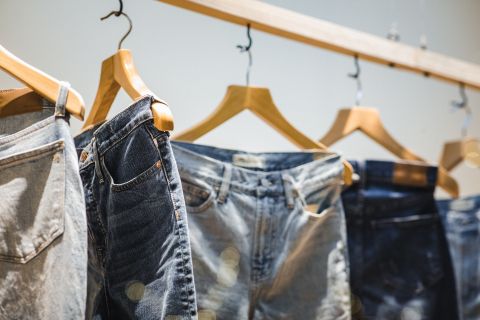 Una collezione di jeans in diverse tonalità è appesa a grucce di legno su una barra di legno. I jeans sono disposti ordinatamente e mostrano una varietà di stili e lavaggi su uno sfondo morbido e neutro.