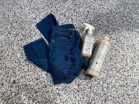 Un paio di jeans blu è appoggiato su uno sfondo screziato grigio e nero. Accanto ai jeans ci sono due prodotti per la pulizia: un flacone spray e un flacone di detersivo, entrambi con etichette minimaliste.