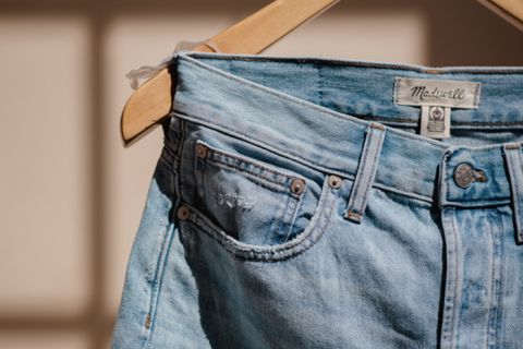 Primo piano di un paio di jeans azzurri appesi a una gruccia di legno. I jeans presentano un'etichetta in pelle con il marchio "Madewell" sulla cintura e sono immersi in una calda luce solare, che mette in risalto la loro texture sbiadita e le cuciture.