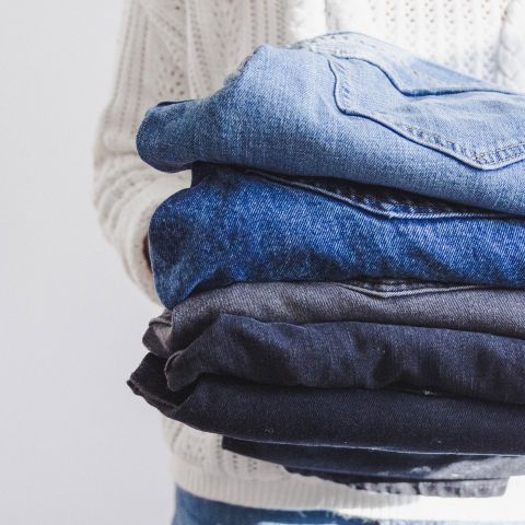 Una persona tiene una pila di jeans piegati. I jeans sono di diverse tonalità di blu e nero. La persona indossa un maglione bianco lavorato a maglia con una trama visibile sullo sfondo.