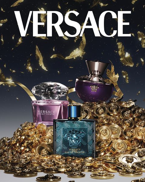 Flaconi di profumi Versace - Bright Crystal, Dylan Purple ed Eros Eau de Toilette - sono circondati da un mucchio di monete d'oro e piume. Il logo Versace è ben visibile in alto su uno sfondo scuro.