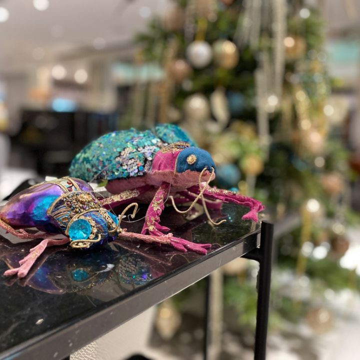 Due sculture di insetti colorate e tempestate di gioielli sono posate su una superficie nera. Gli insetti sono adornati con tessuti e perline luminose nei toni del rosa, del blu e del viola. Sullo sfondo, un albero di Natale decorato è sfocato.