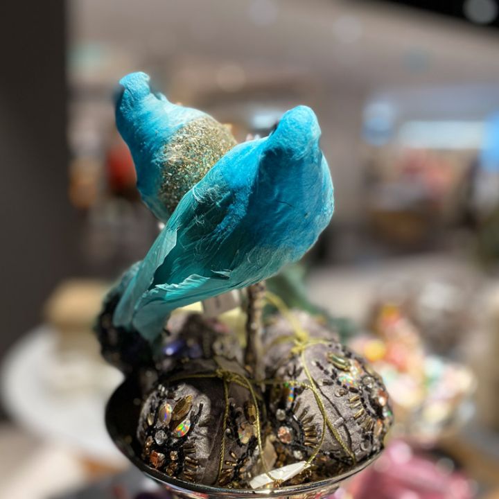 Due uccelli decorativi blu sono appollaiati su un ramo ornato di strass e glitter. Lo sfondo è leggermente sfocato, il che esalta i colori vivaci e i dettagli intricati degli uccelli.