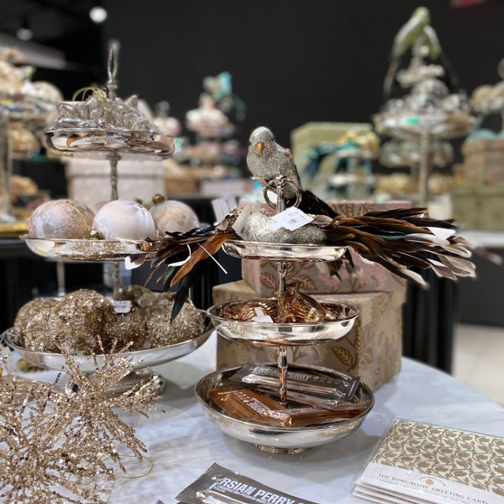 Una mostra di decorazioni natalizie con vassoi argentati a più livelli adornati con ornamenti scintillanti, una figura di uccello con piume e oggetti decorativi. Sullo sfondo si possono vedere altre decorazioni festive per la tavola.