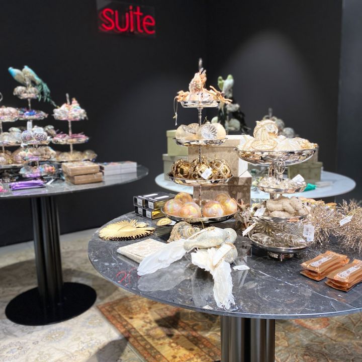 In una vetrina si vedono due tavoli rotondi con vassoi decorati a più livelli, sui quali sono disposti vari oggetti decorativi come ornamenti e piume. La parete di fondo è scura e riporta la scritta "Suite" in neon rosso. Sul pavimento si intravede parzialmente un tappeto a motivi.