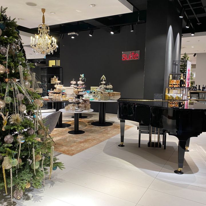 Un lussuoso interno di negozio con un albero di Natale decorato, un pianoforte a coda nero e un'esposizione di torte e pasticcini elaborati su eleganti supporti. L'ambiente è festoso e raffinato, con un lampadario e decorazioni di stile.