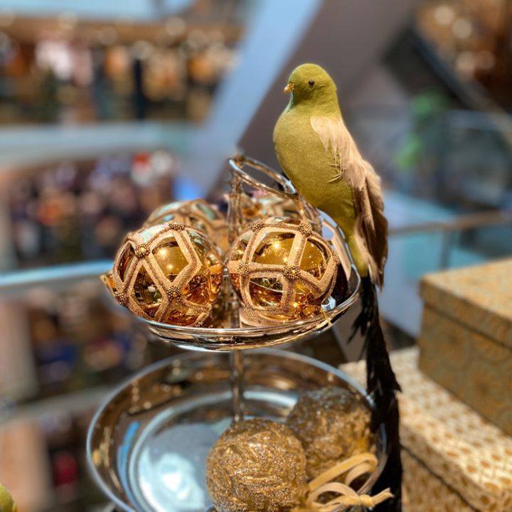 Un uccello decorativo verde è appollaiato su un vassoio con ornamenti dorati e scintillanti in una vetrina. Lo sfondo mostra una vista sfocata di un centro commerciale a più piani.