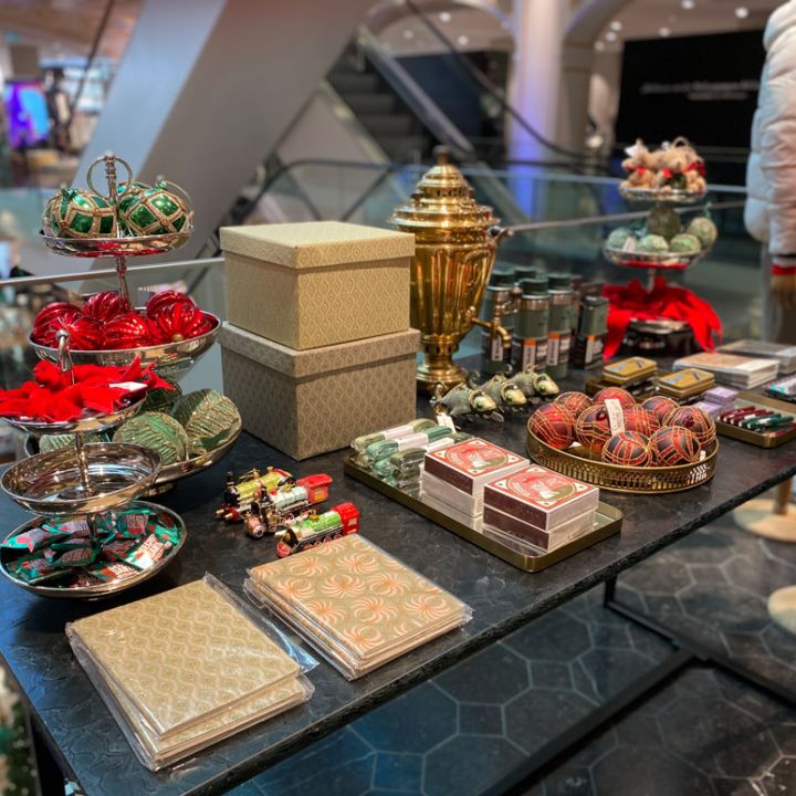 Su un tavolo espositivo in un negozio sono esposte varie decorazioni e regali natalizi, tra cui ornamenti natalizi, scatole confezionate e dolciumi. Sullo sfondo si vedono una scala mobile e manichini con abiti invernali.