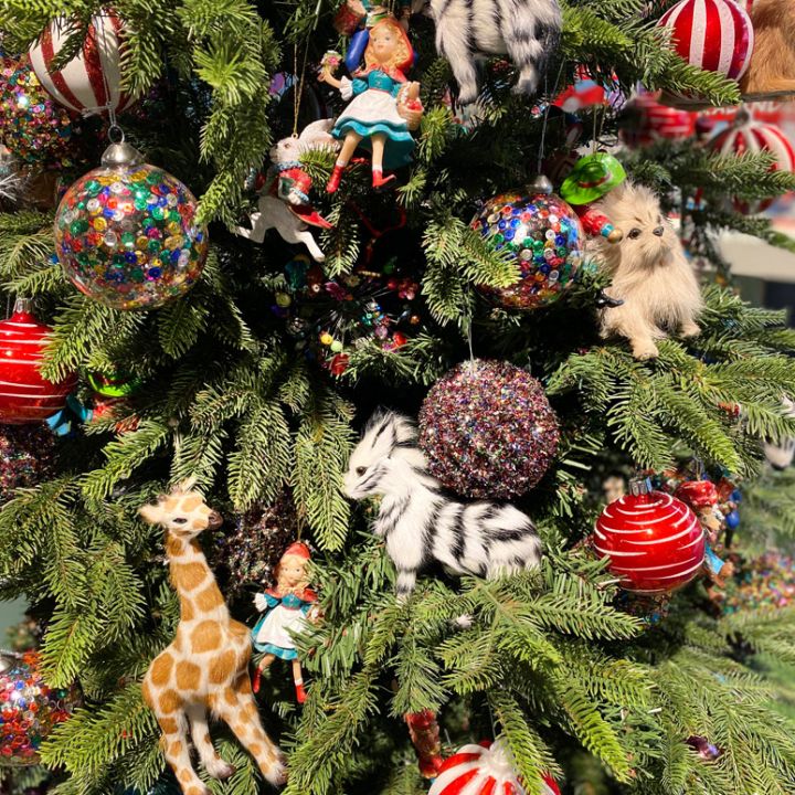 Un albero di Natale decorato con ornamenti colorati, tra cui palline decorate, peluche come una giraffa e una zebra, e piccole bambole. L'albero è rigoglioso di rami verdi e decorazioni colorate, creando una festosa scena natalizia.