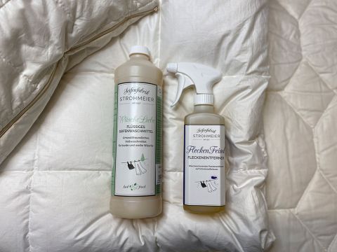 Su una superficie bianca trapuntata ci sono due flaconi di detergenti. Uno è un detersivo liquido in un flacone alto, l'altro è uno spray smacchiatore in un flacone più piccolo. Entrambi hanno etichette bianche con testo in tedesco.