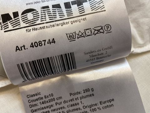 Primo piano di un'etichetta in tessuto su un piumone con simboli e istruzioni per la cura. Contiene dettagli del prodotto come "Classic", dimensioni "140x200 cm", peso "250 g" e menziona materiali come "100% cotone". Ulteriori testi includono un codice a barre e informazioni sull'azienda.