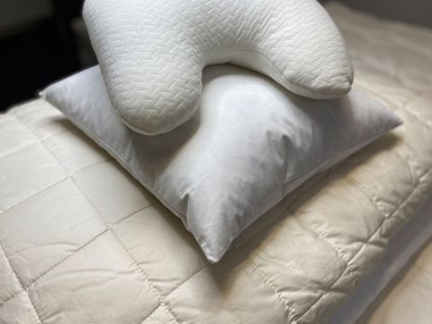 Un cuscino bianco a forma di U è appoggiato su un normale cuscino bianco, il tutto su un copriletto trapuntato beige. La scena trasmette una sensazione di comfort e relax.