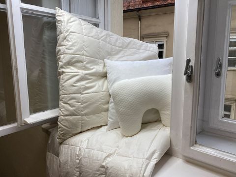 Angolo accogliente della finestra con soffici cuscini bianchi e una trapunta su una sporgenza. Due finestre aperte offrono una vista su un edificio dall'esterno. I cuscini includono un grande cuscino quadrato, uno rettangolare più piccolo e un cuscino da viaggio a forma di U.