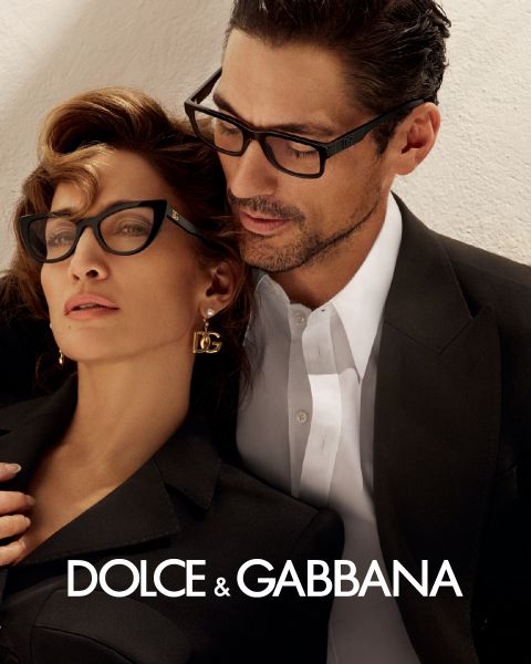 Una donna e un uomo con occhiali eleganti sono appoggiati vicini. Lei indossa orecchini DG e un abito nero, lui indossa un abito nero con una camicia bianca. In basso si vede il logo di Dolce & Gabbana.