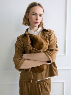 Una donna con un bob biondo, che indossa un outfit marrone, sta in piedi con le braccia incrociate davanti a un muro bianco, tenendo in mano una grande borsa di camoscio marrone.