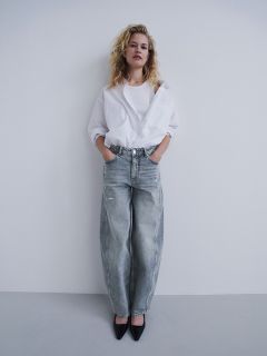 Una donna in camicia bianca e jeans grigi larghi sta in piedi con le mani in tasca davanti a un muro bianco, indossa décolleté nere e guarda la fotocamera.