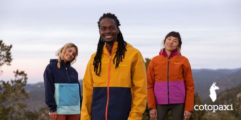 Tre persone in giacche outdoor colorate si trovano in un paesaggio naturale con alberi e montagne. Il cielo è coperto. In basso a destra si vede il logo del marchio "Cotopaxi".