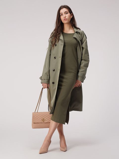 Una donna in un abito verde oliva e un trench coat abbinato tiene in mano una borsa trapuntata beige con tracolla a catena. Indossa tacchi alti beige, è in piedi davanti a uno sfondo grigio semplice e guarda con sicurezza la fotocamera.