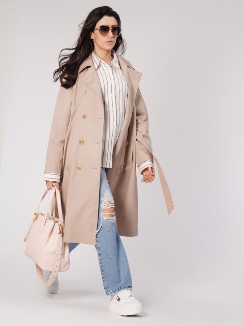 Una persona con capelli lunghi, occhiali da sole, trench beige, camicia a righe, jeans strappati e scarpe da ginnastica bianche porta una borsa luminosa. Cammina con sicurezza davanti a uno sfondo neutro.