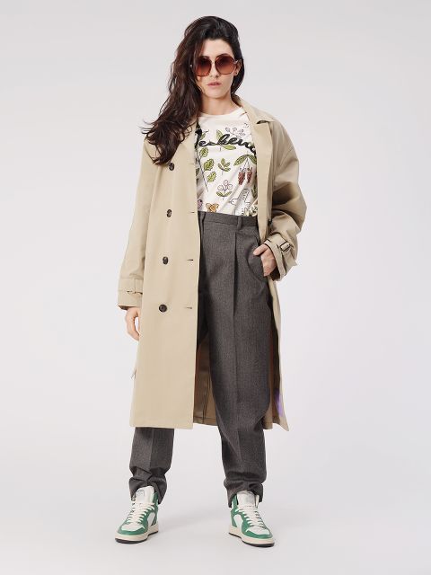 Una persona con un trench beige, una camicia a motivi, pantaloni grigi, scarpe da ginnastica verdi e bianche e occhiali da sole posa con sicurezza davanti a uno sfondo semplice. I suoi capelli sono lunghi e acconciati in morbide onde.