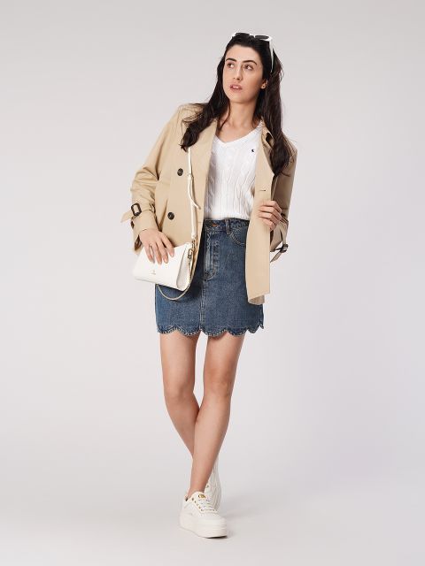 Una donna è in piedi davanti a uno sfondo semplice. Indossa un trench beige, un top bianco e una gonna di jeans. Ha una piccola borsa a tracolla bianca e scarpe da ginnastica bianche. I suoi lunghi capelli sono sciolti e indossa un paio di occhiali da sole sulla testa.
