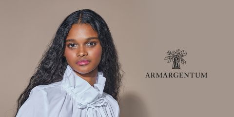 Una donna con lunghi capelli ondulati indossa una camicetta bianca con colletto alto. È in piedi davanti a uno sfondo beige, sul cui lato destro si trova il logo "ARMARGENTUM" con un motivo floreale.