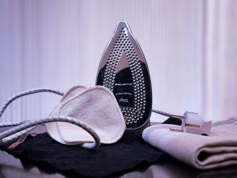 Un ferro da stiro è posizionato verticalmente su una superficie riflettente, circondato da una camicia stirata, una mascherina di stoffa e un cavo. Lo sfondo è sfocato, evidenziando l'ambiente domestico.