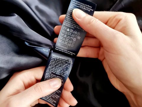 Le mani tengono un'etichetta scura di un indumento con istruzioni per la cura, inclusi simboli per il lavaggio, la candeggina, l'asciugatura e la stiratura. L'etichetta ha testo bianco su sfondo scuro e l'indumento sembra essere fatto di un tessuto setoso.