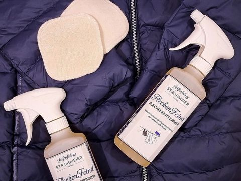Su una giacca trapuntata blu scuro sono posizionate due bottiglie di spray detergente con la scritta "FleckenFeind", accanto a due dischetti riutilizzabili per la pulizia color beige. Gli erogatori spray sono bianchi e la disposizione suggerisce un'attenzione alla cura del bucato.
