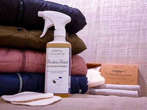 Su una pila di giacche piegate e colorate si trova un flacone spray con l'etichetta "Flecken-Feind Smacchiatore". Accanto ci sono campioni di tessuto e una scatola. Lo sfondo presenta una superficie strutturata.