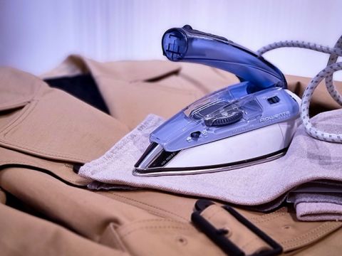 Un ferro da stiro blu e argento è appoggiato su una giacca beige, che si trova su una superficie piana. Un cavo arrotolato parte dal ferro da stiro. La giacca ha un passante per cintura e una fibbia.